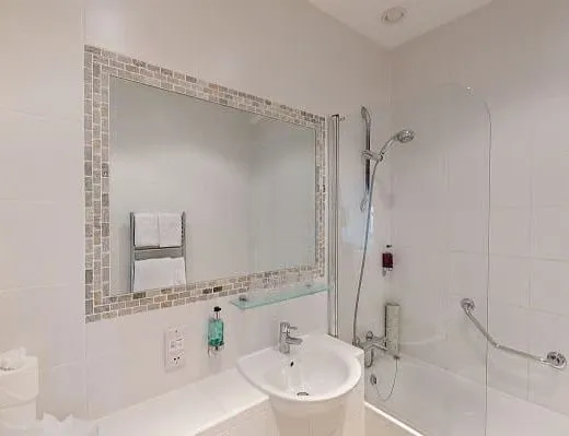 Bright family bathroom with white tiles, a bath and shower, pedestal sink, and a large mirror with mosaic trim