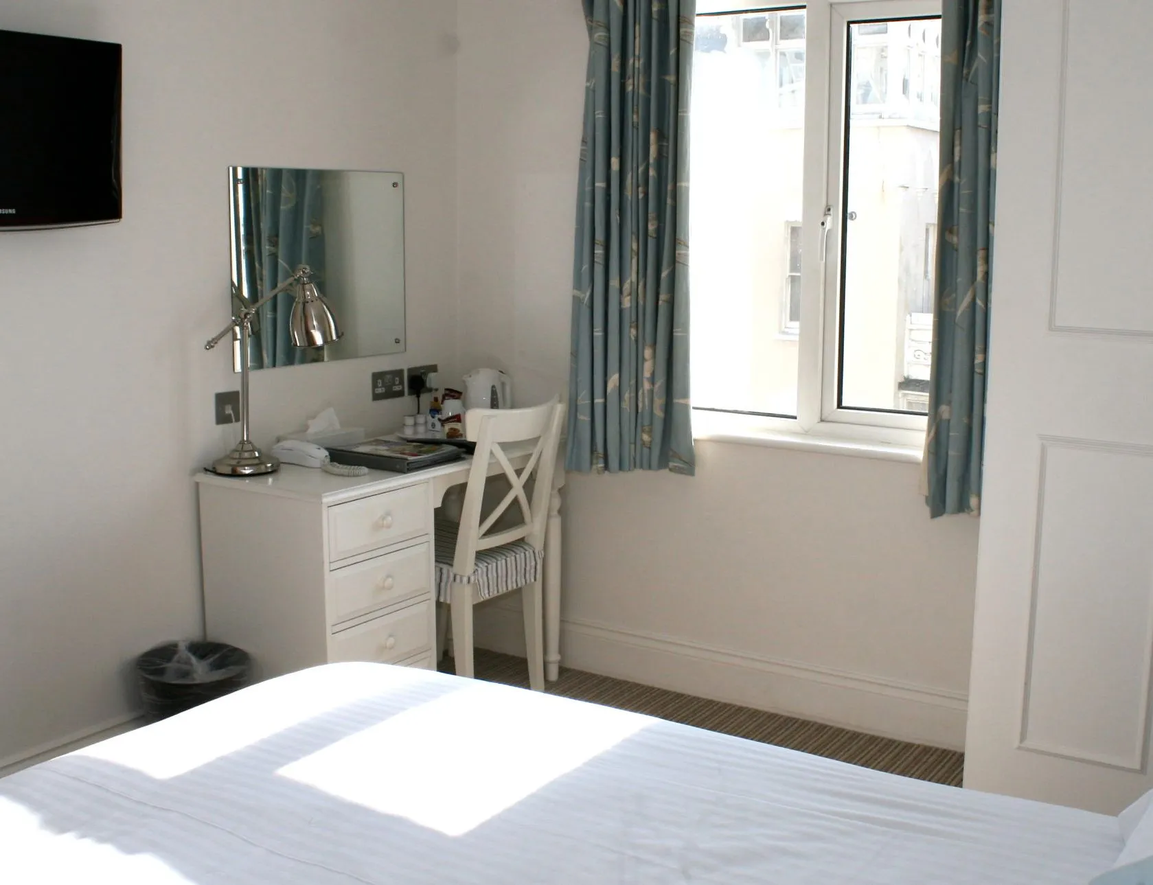 Bright single bedroom with a neatly made bed, wall-mounted television, dressing table and chair by a window with blue curtains.