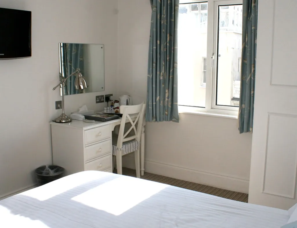 Bright single bedroom with a neatly made bed, wall-mounted television, dressing table and chair by a window with blue curtains.