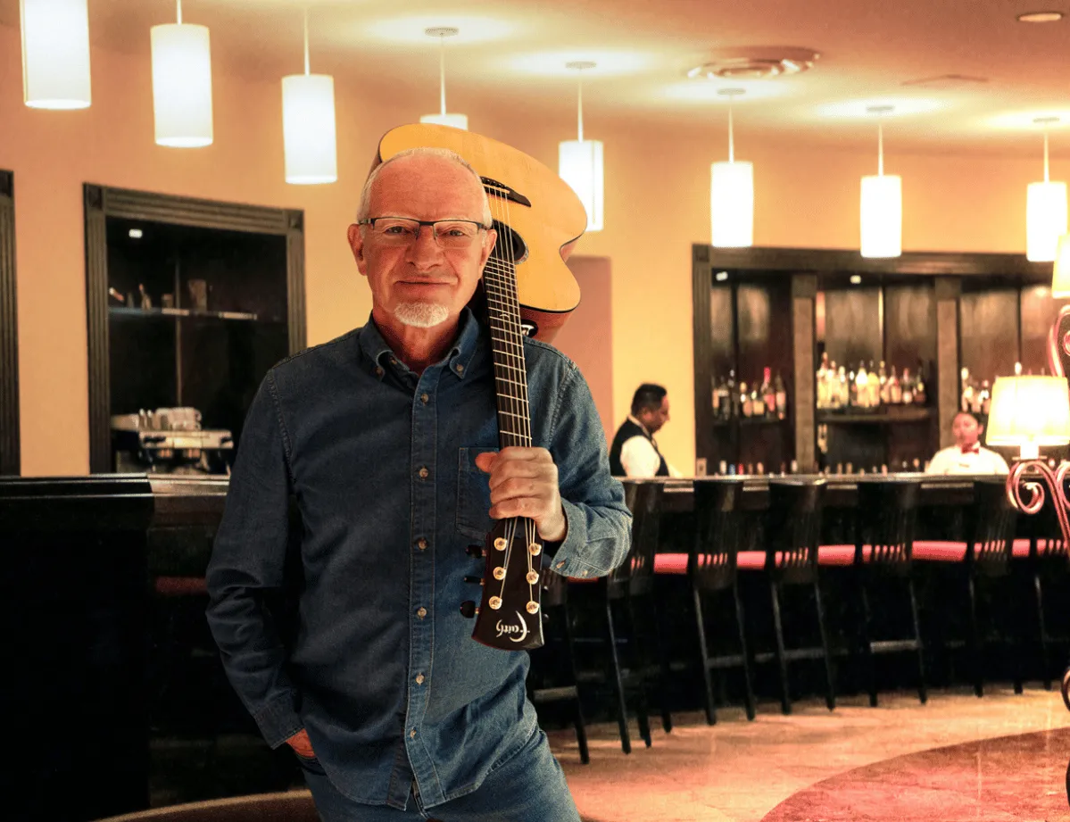 Older man holding an acoustic guitar in a warmly lit bar or lounge