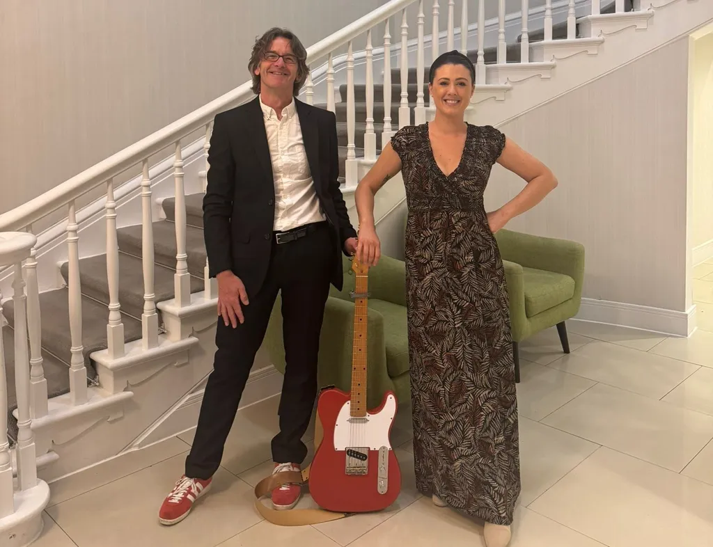 Natasha Wilson duo standing beside a red electric guitar in an elegant interior with a sweeping staircase