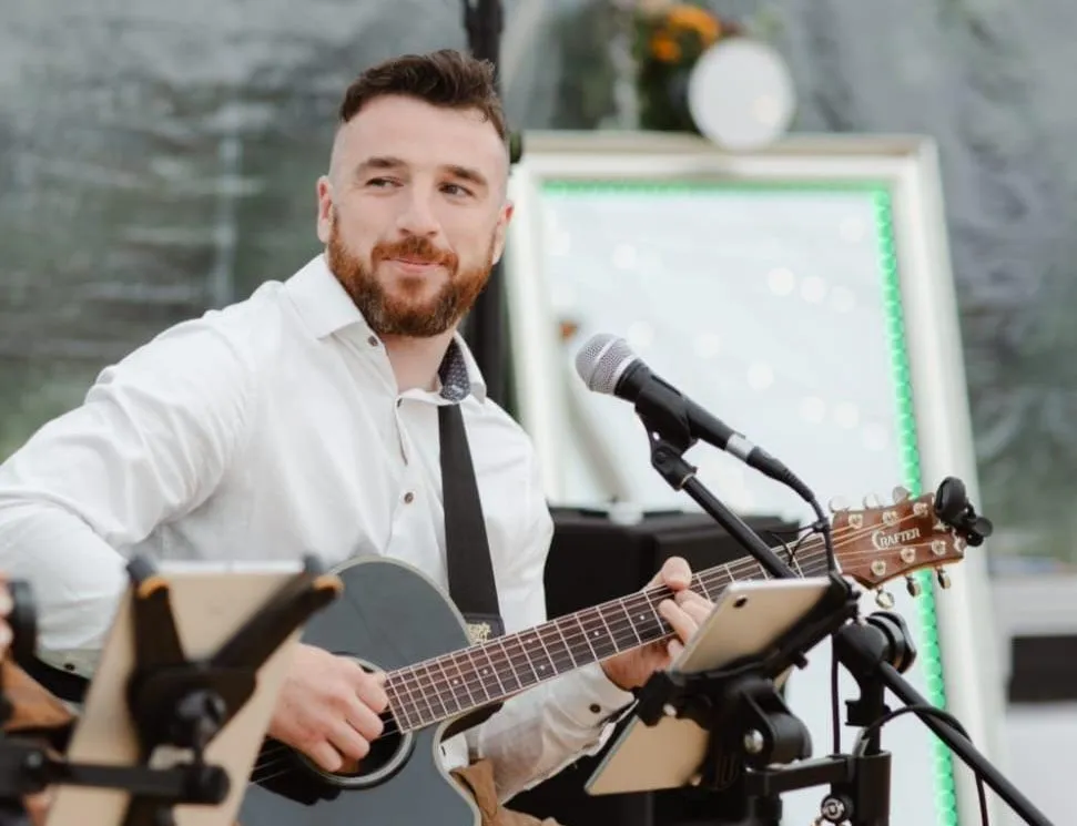 Bearded male musician in a white shirt playing an acoustic guitar and singing into a microphone