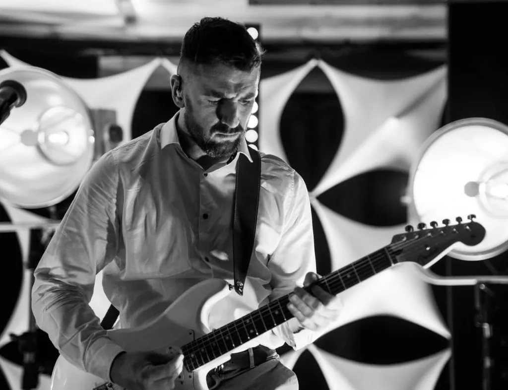 Black and white photo of a guitarist performing on stage with an electric guitar and amplifier behind him.