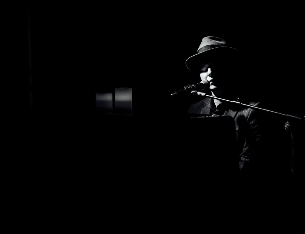 Black and white portrait of a singer in a hat performing at a microphone in low light.