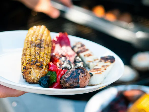 Plate of grilled vegetables with charred sweetcorn, peppers, onion and halloumi by a barbecue