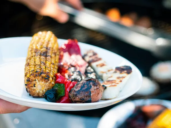 Plate of grilled vegetables with charred sweetcorn, peppers, onion and halloumi by a barbecue