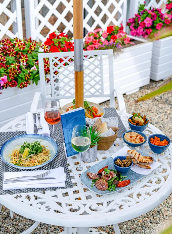 Outdoor dining table at Ommaroo food terrace set with pasta, charcuterie, bread, dips and glasses of wine beside colourful flower planters
