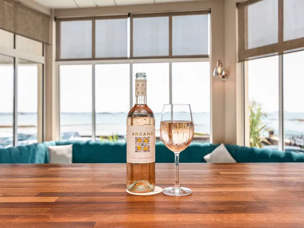 Bottle of rosé wine and a glass on a wooden bar table with sea views through large windows