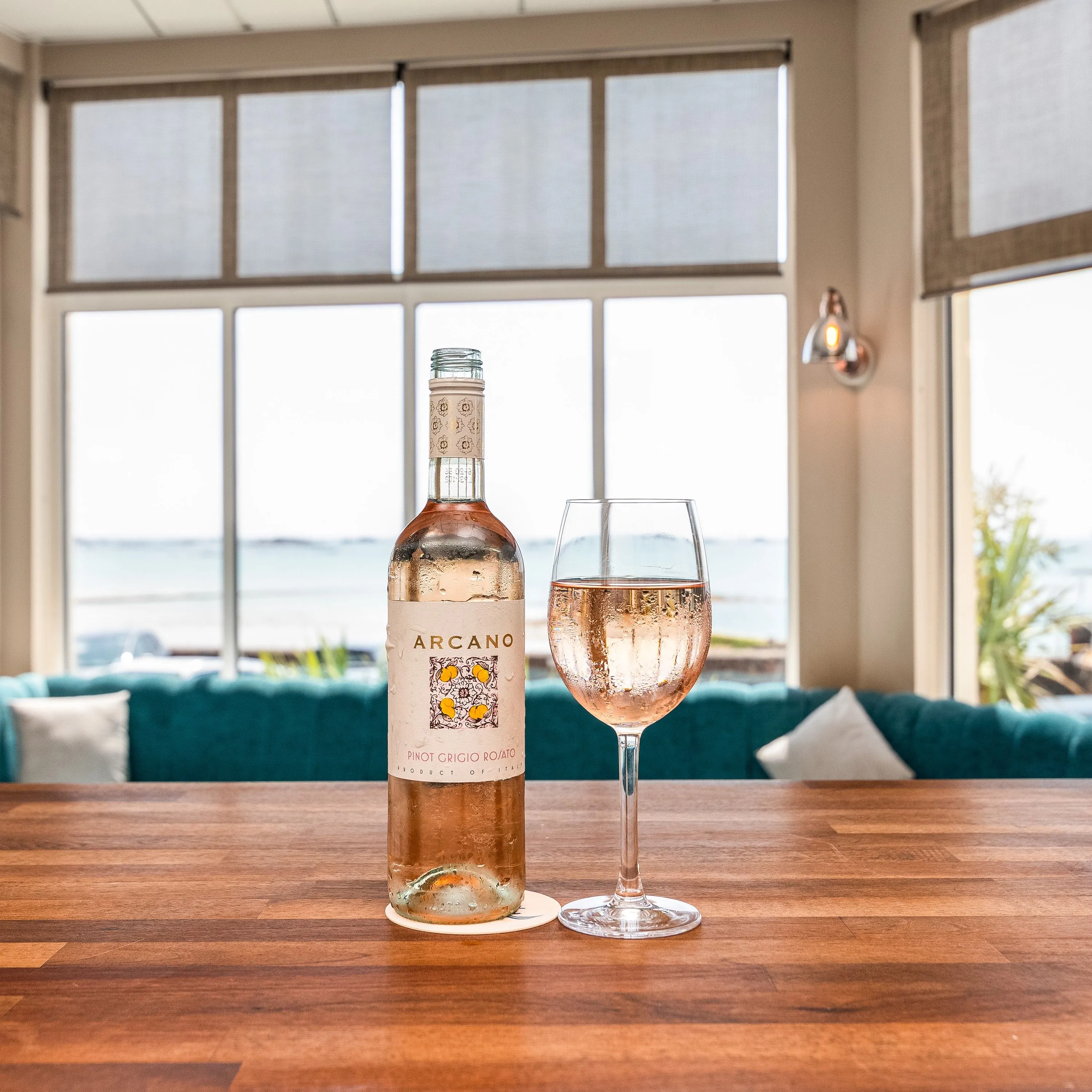 Bottle of rosé wine and a glass on a wooden bar table with sea views through large windows