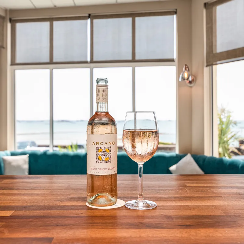 Bottle of rosé wine and a glass on a wooden bar table with sea views through large windows