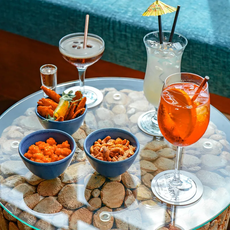 Close-up of cocktails and bar snacks on a glass-topped table beside a pool at Coast Bar.