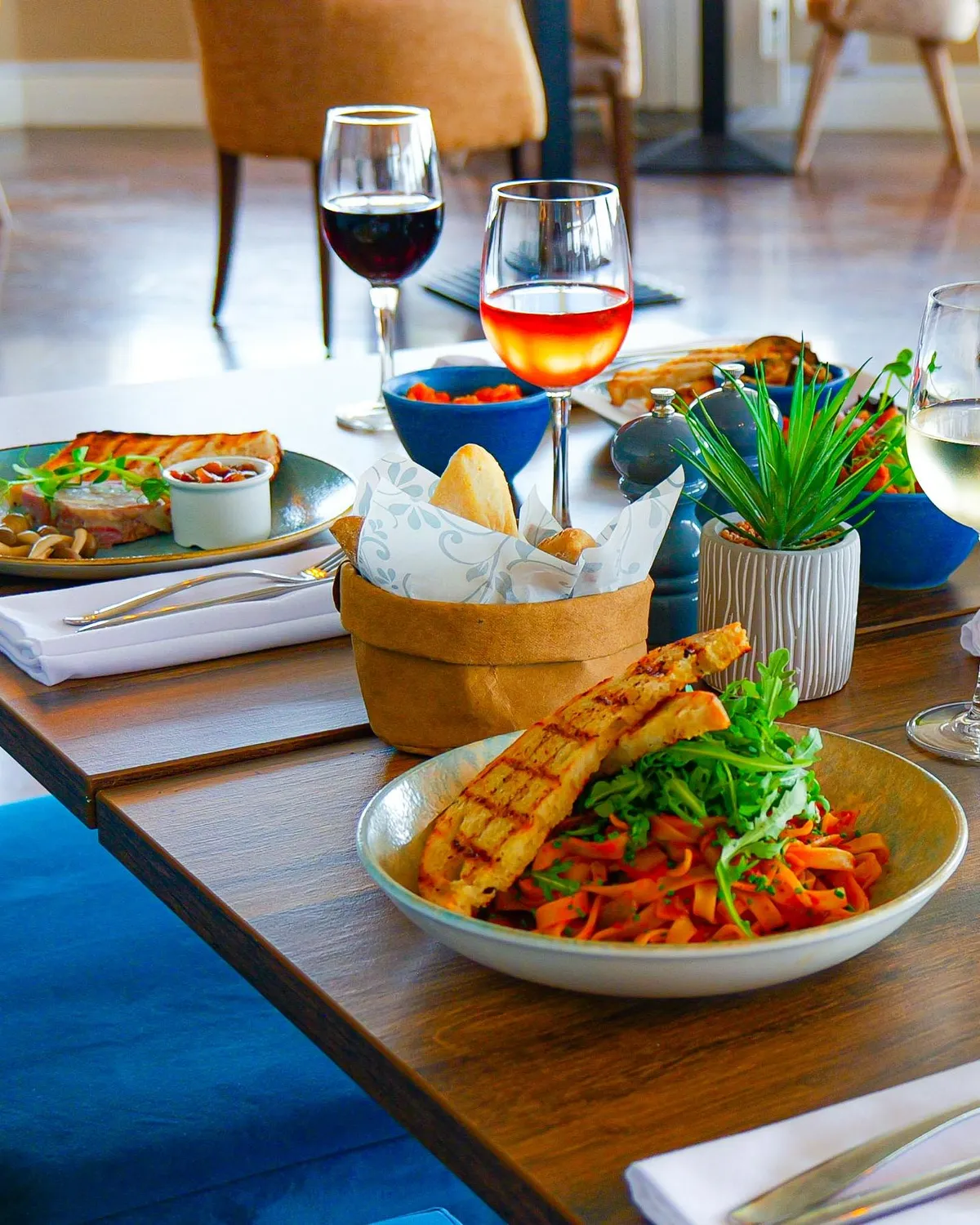 Close-up of a Coast Bistro table in the bar area set with pasta dishes, bread, crisps and glasses of red, rosé and white wine.