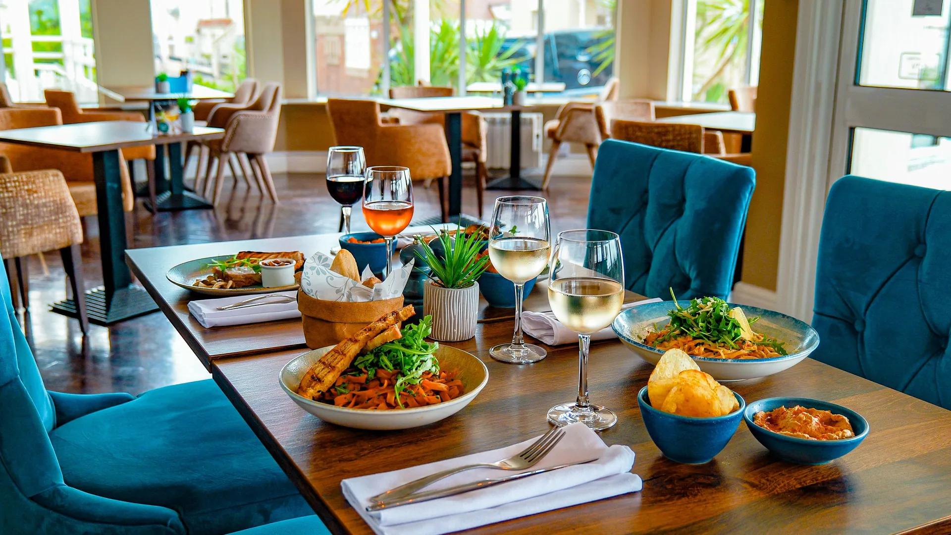 Dining table set with pasta dishes, bread, crisps and glasses of wine in the bright bar area at Coast Bistro.