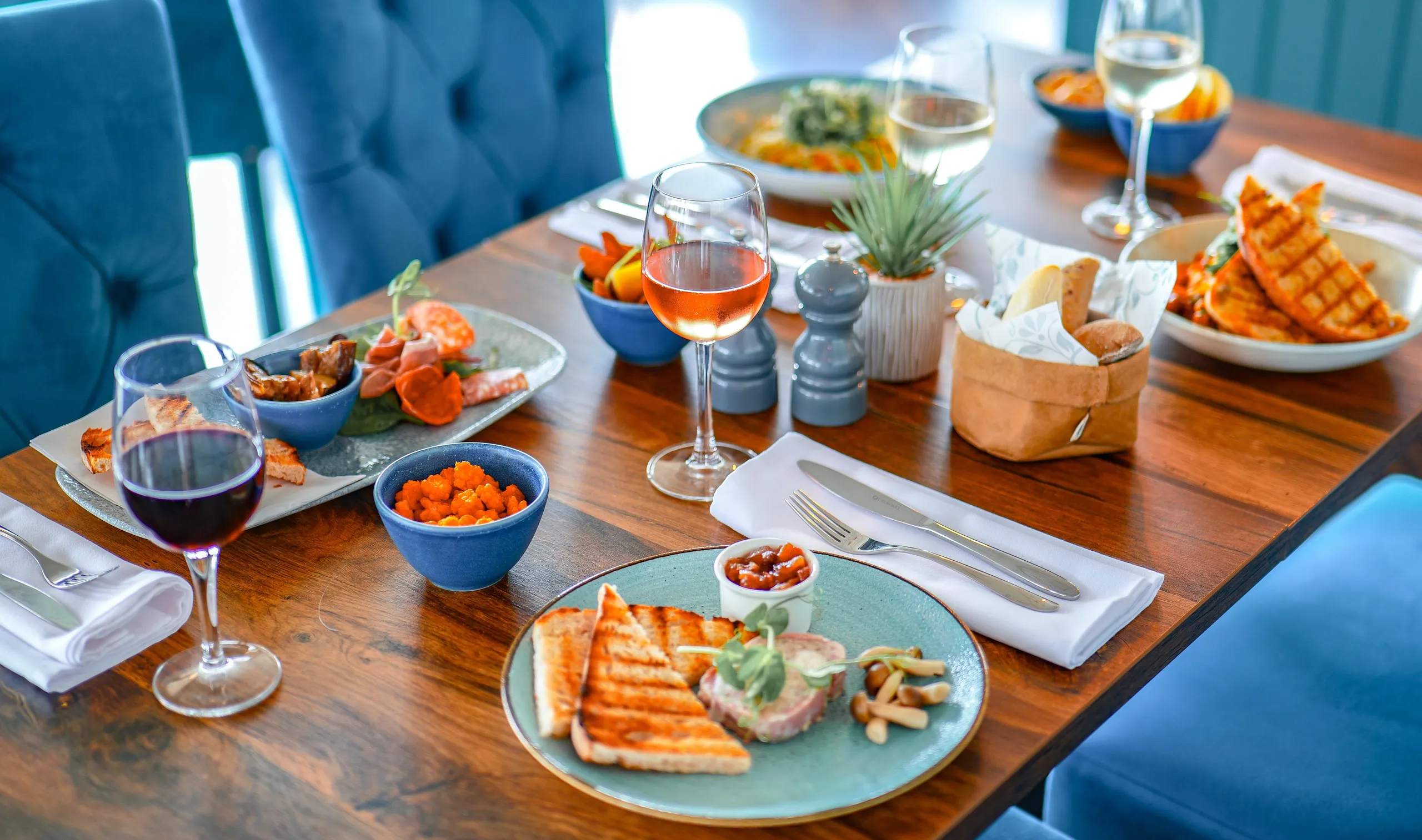 Close-up of a Coast Bistro table set with sharing plates, grilled sandwiches, wine and bread on a wooden table.