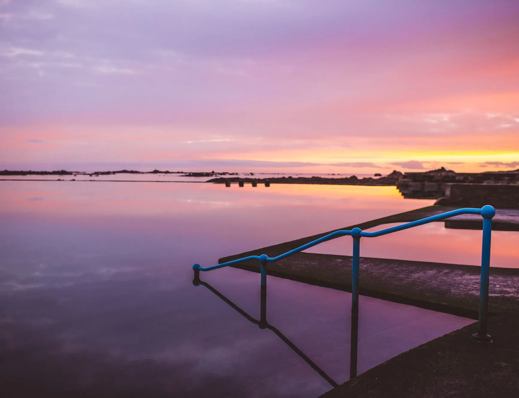 Still lido pool at sunset with blue handrails and a pink and orange sky reflected in the water