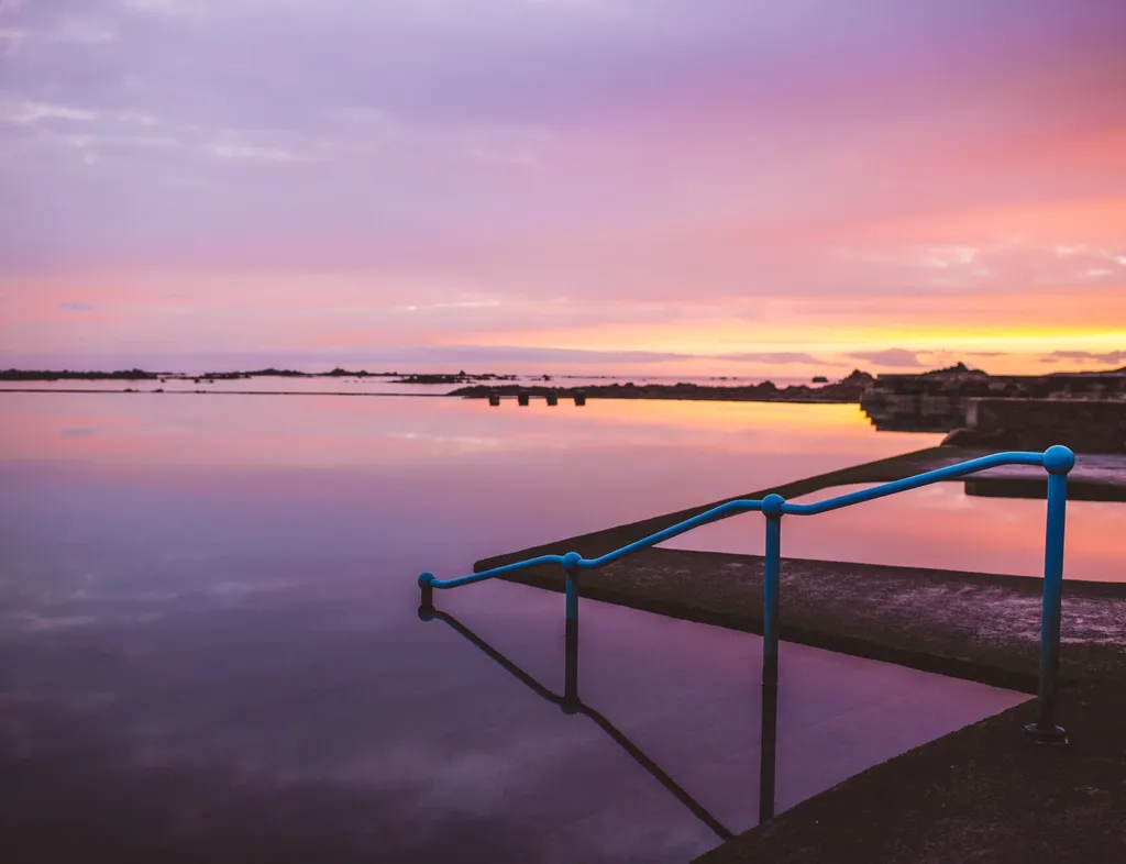 Still lido pool at sunset with blue handrails and a pink and orange sky reflected in the water