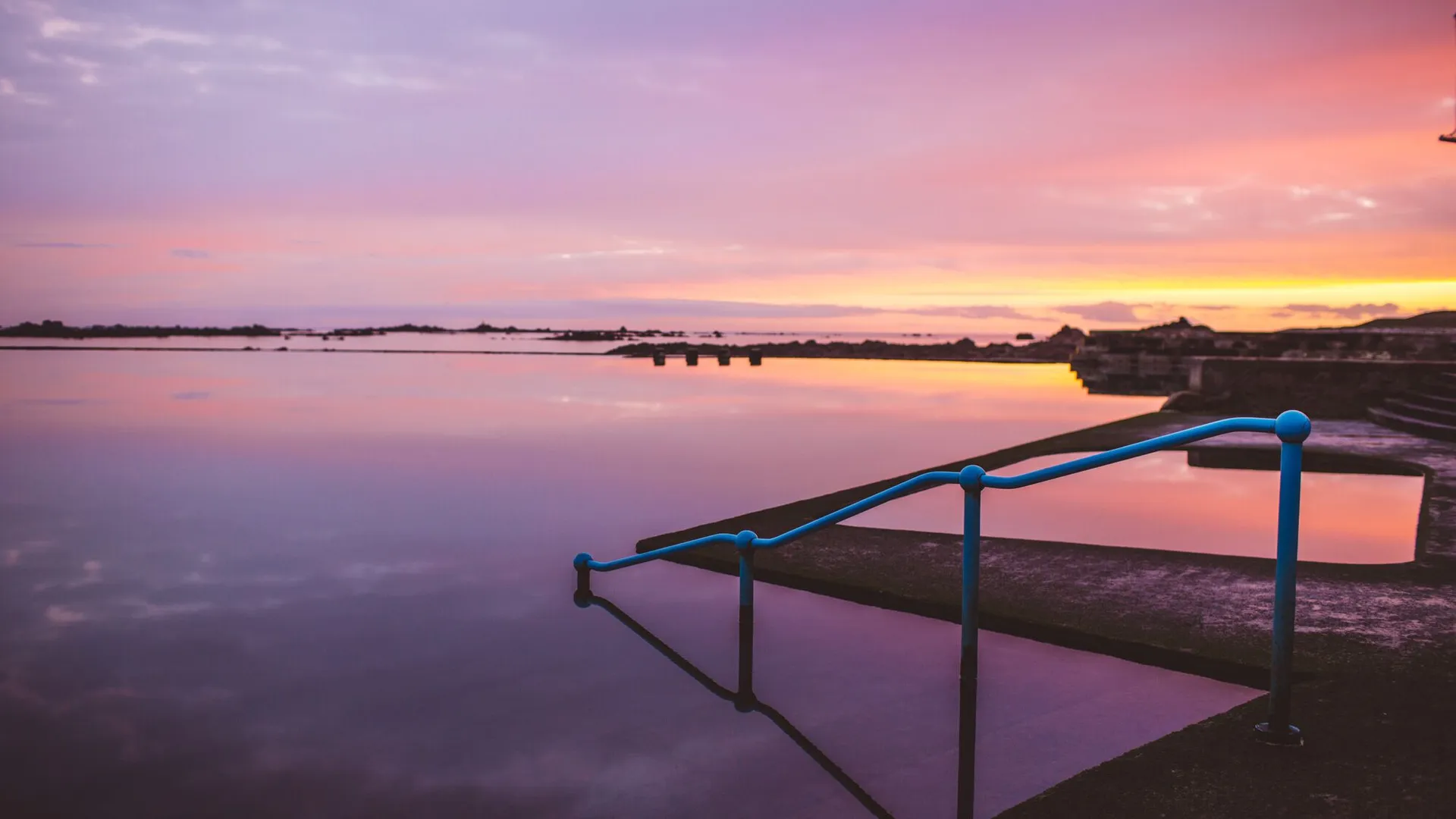 Still lido pool at sunset with blue handrails and a pink and orange sky reflected in the water