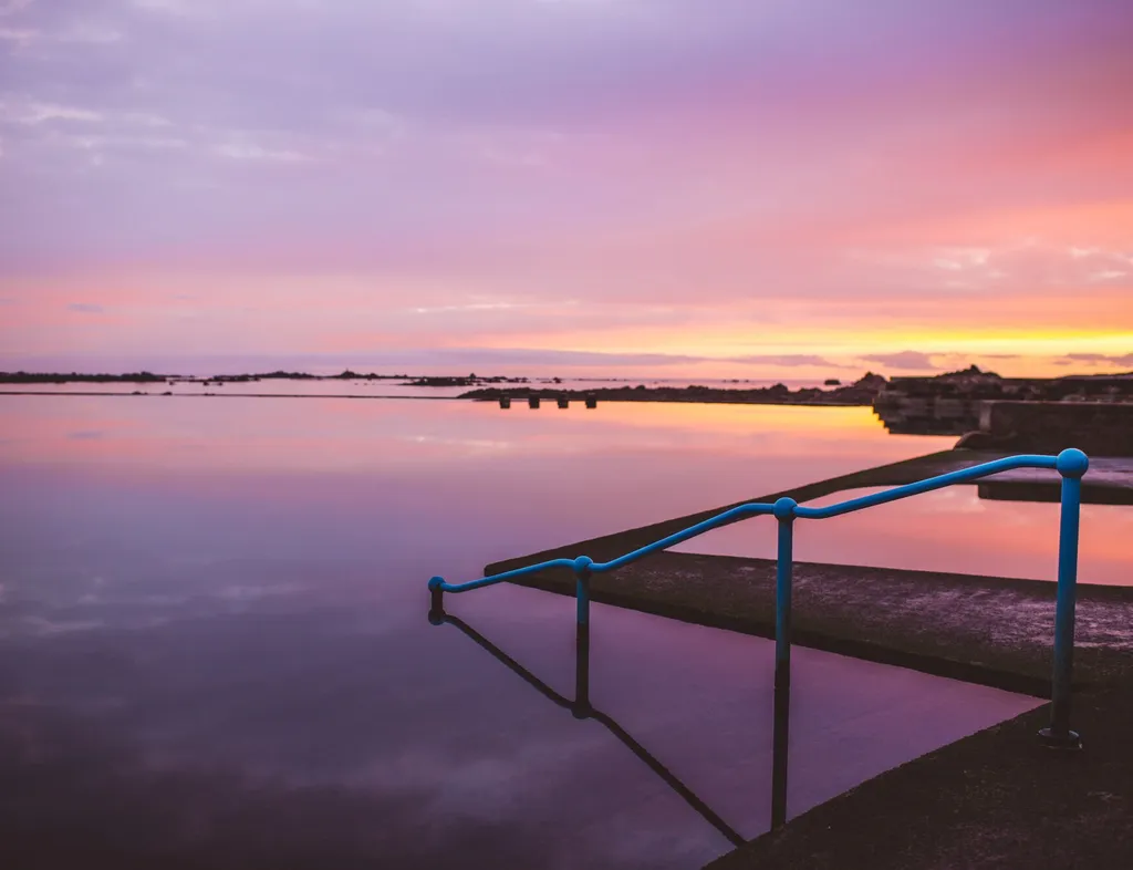 Still lido pool at sunset with blue handrails and a pink and orange sky reflected in the water