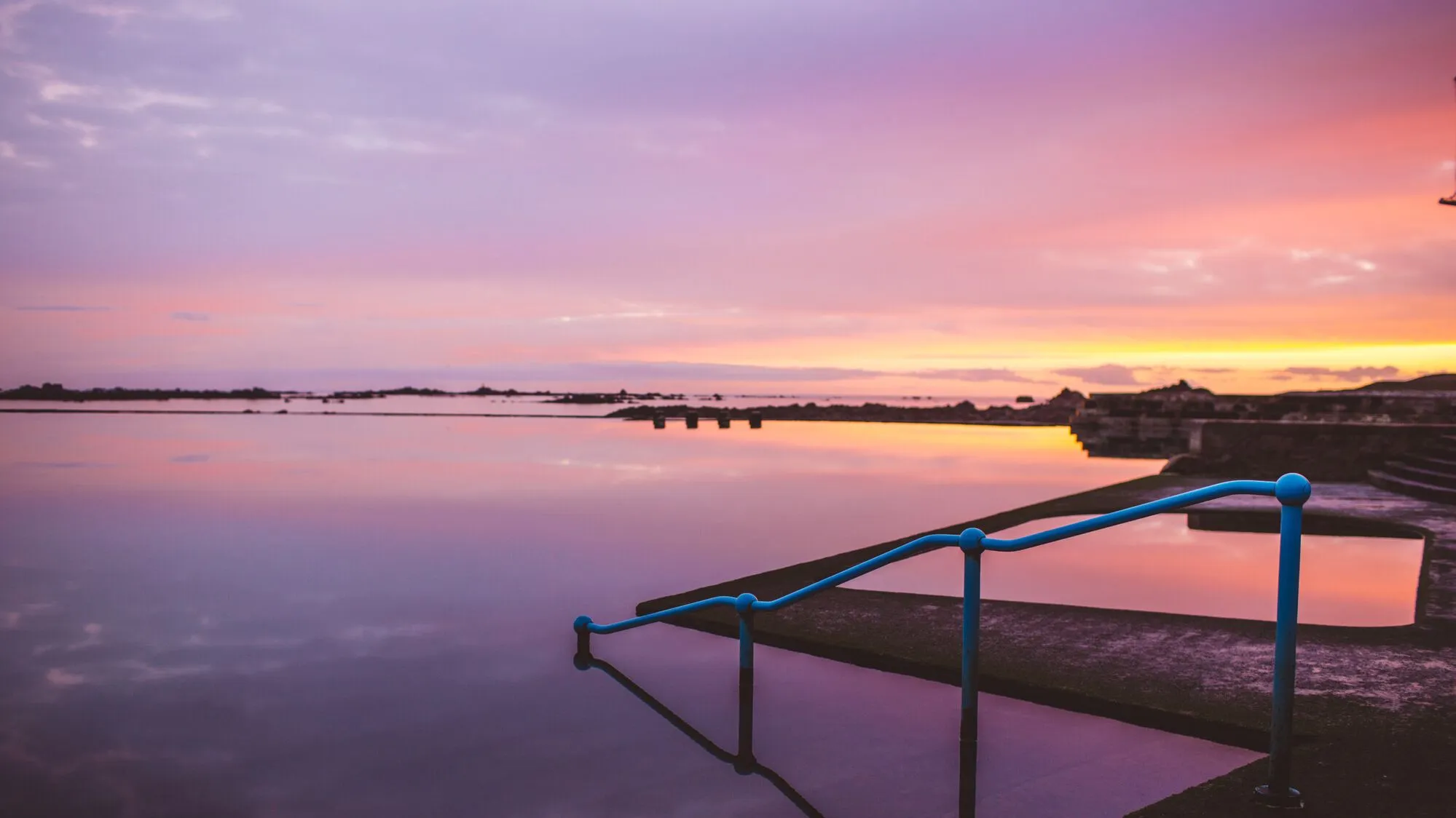 Still lido pool at sunset with blue handrails and a pink and orange sky reflected in the water