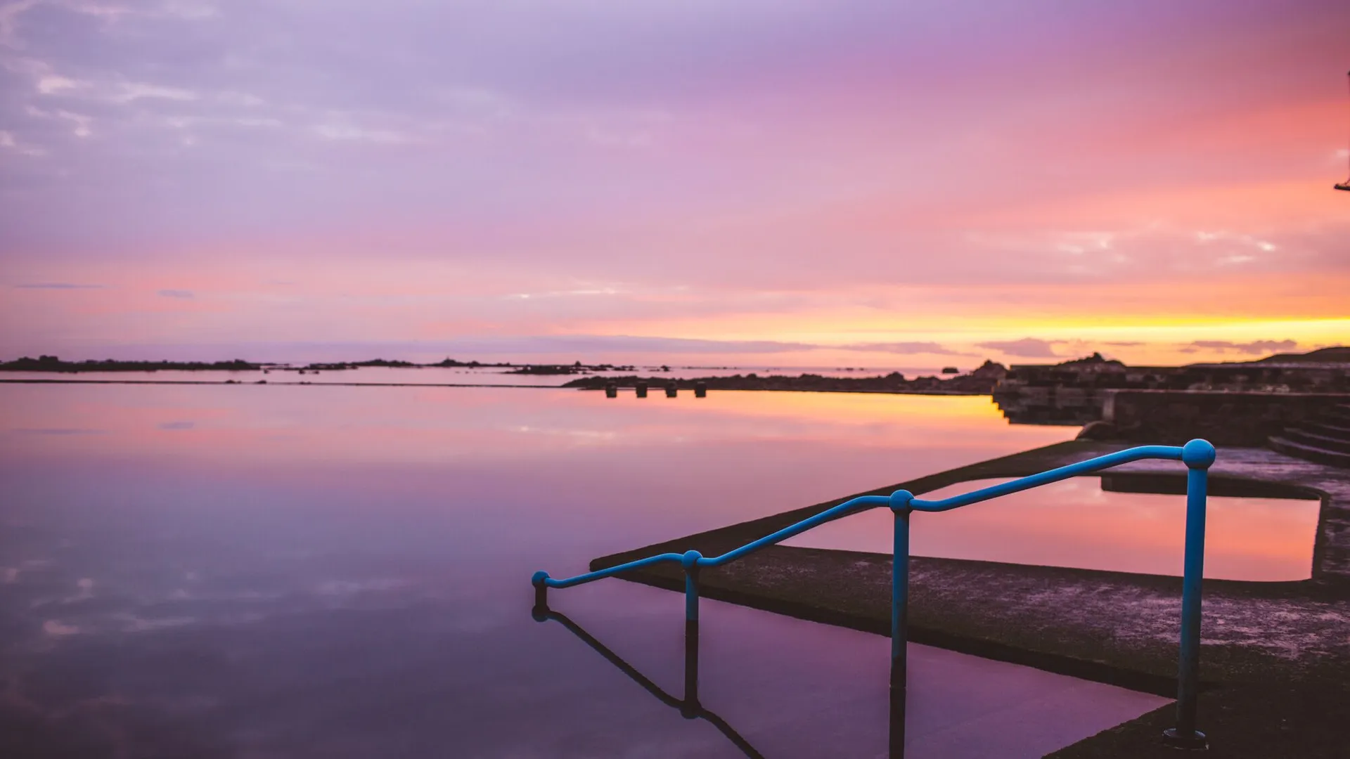 Still lido pool at sunset with blue handrails and a pink and orange sky reflected in the water