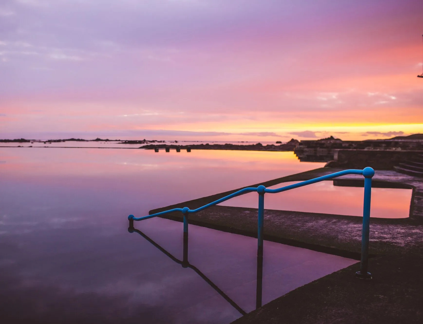 Still lido pool at sunset with blue handrails and a pink and orange sky reflected in the water