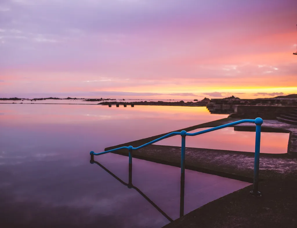 Still lido pool at sunset with blue handrails and a pink and orange sky reflected in the water