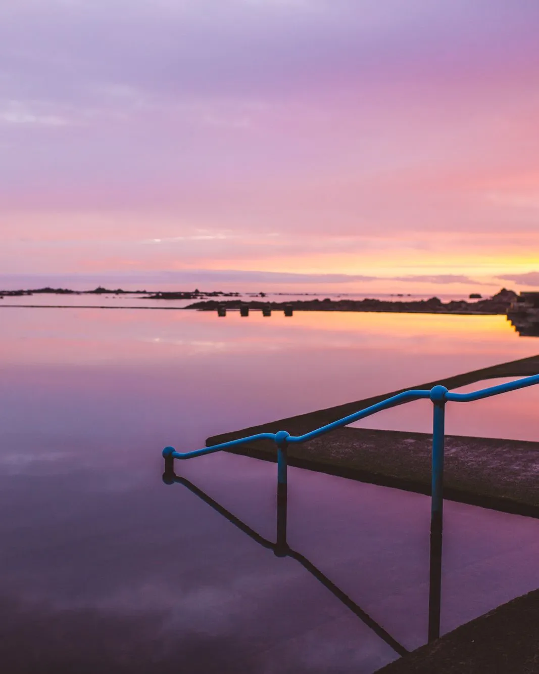 Still lido pool at sunset with blue handrails and a pink and orange sky reflected in the water