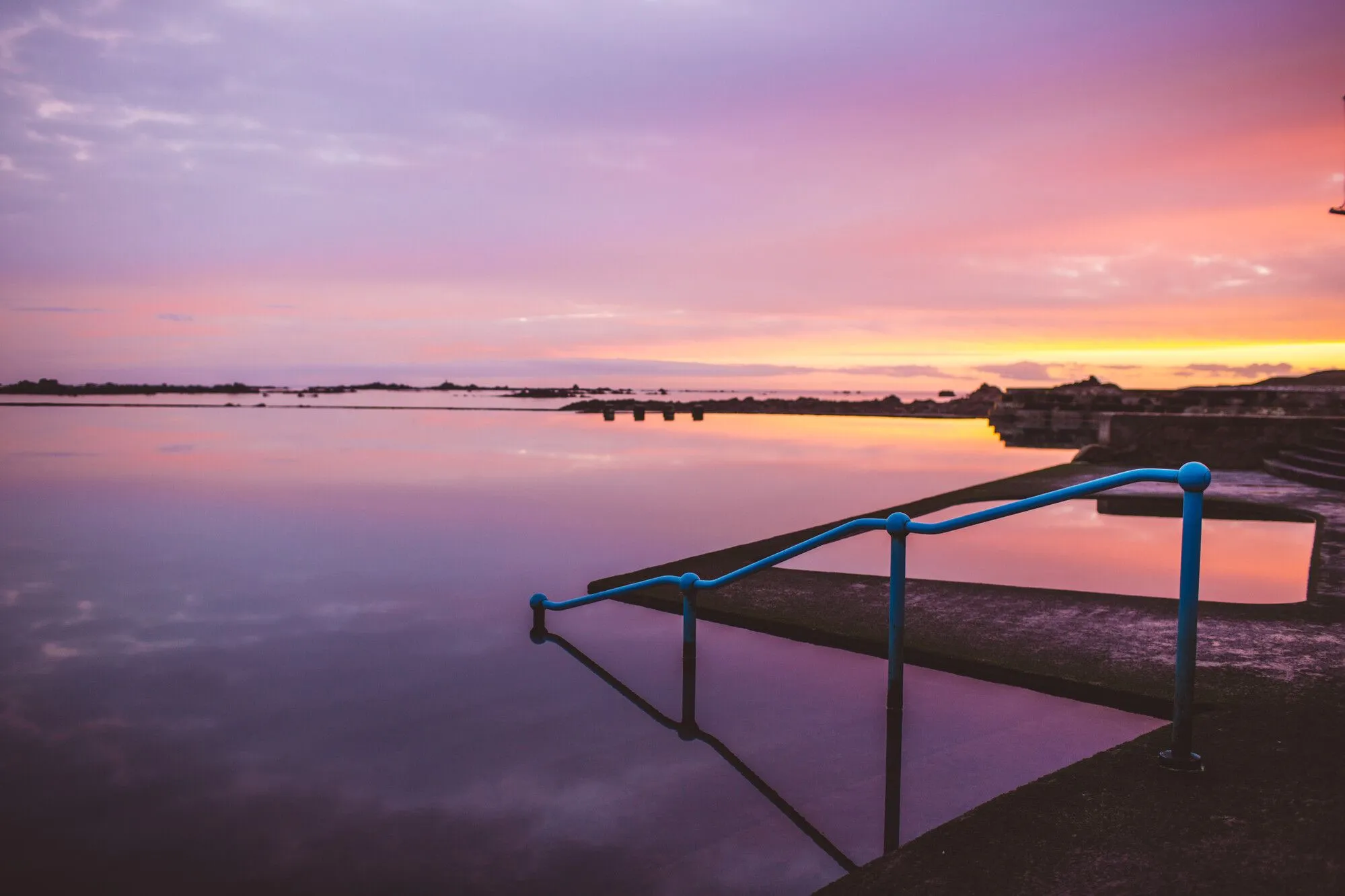 Still lido pool at sunset with blue handrails and a pink and orange sky reflected in the water