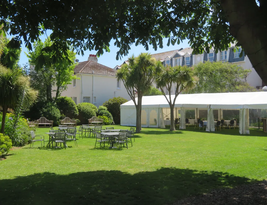 White marquee set up on a sunny garden lawn with outdoor tables and chairs beneath mature trees