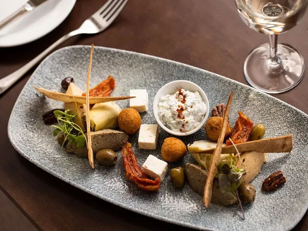 A plated mezze selection with feta, olives, artichokes, sun-dried tomatoes, crisp flatbread and a herb dip on a restaurant table.