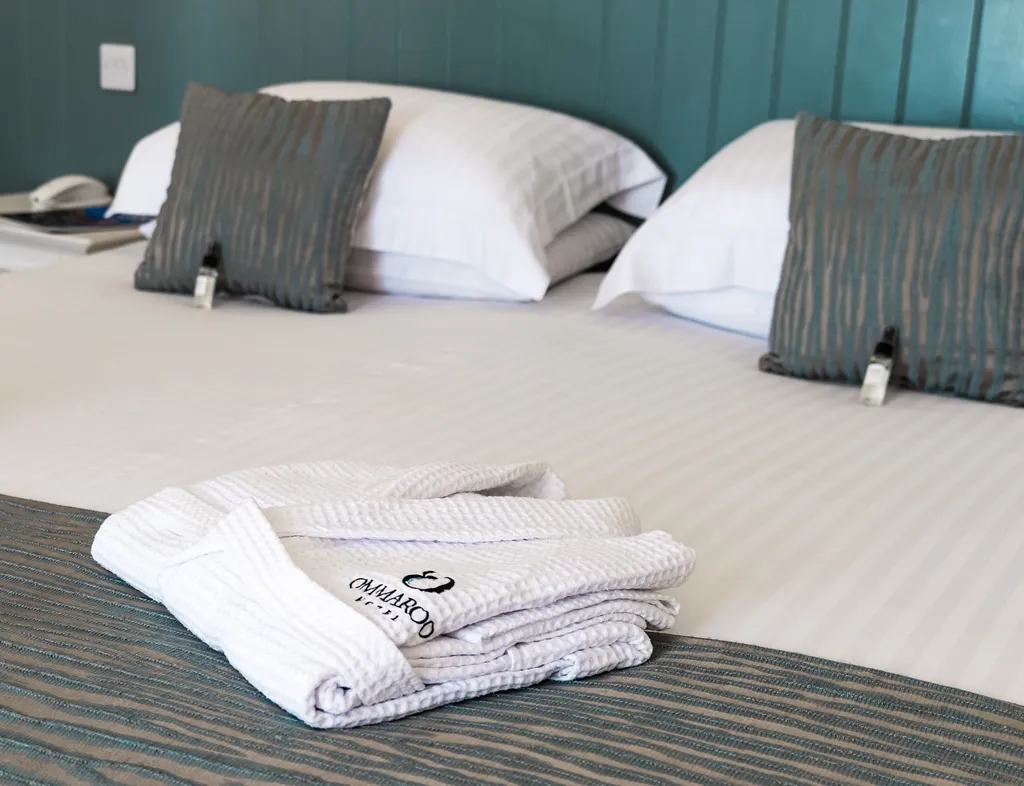 Spacious hotel bed with crisp white linen, teal cushions and folded bathrobes in a seaview balcony room