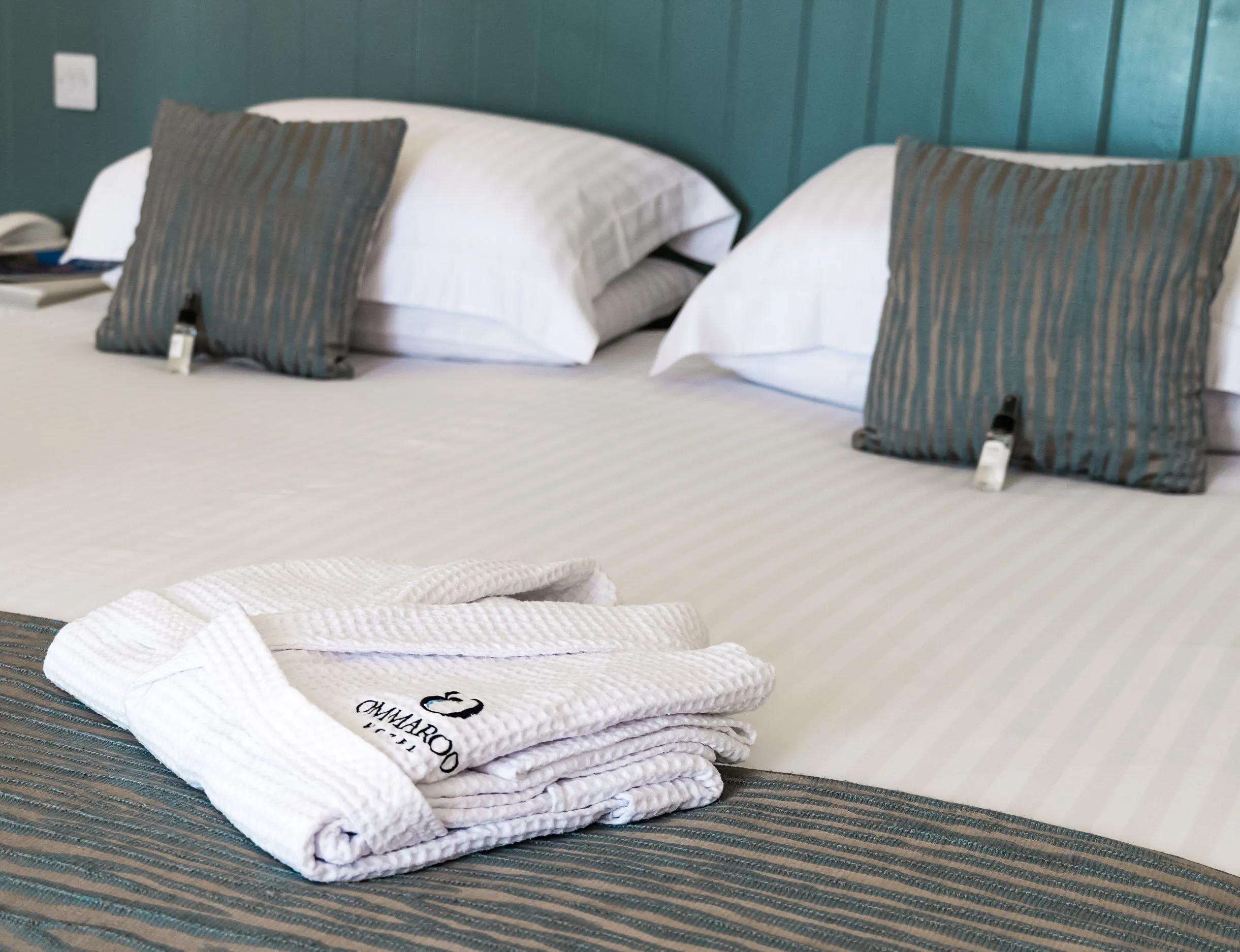 Spacious hotel bed with crisp white linen, teal cushions and folded bathrobes in a seaview balcony room