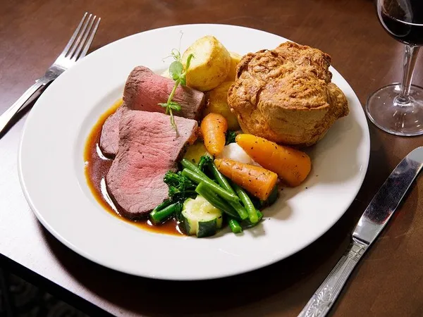 Plate of Sunday roast with sliced beef, Yorkshire pudding, roast potatoes, carrots, green vegetables and gravy, served with a glass of red wine