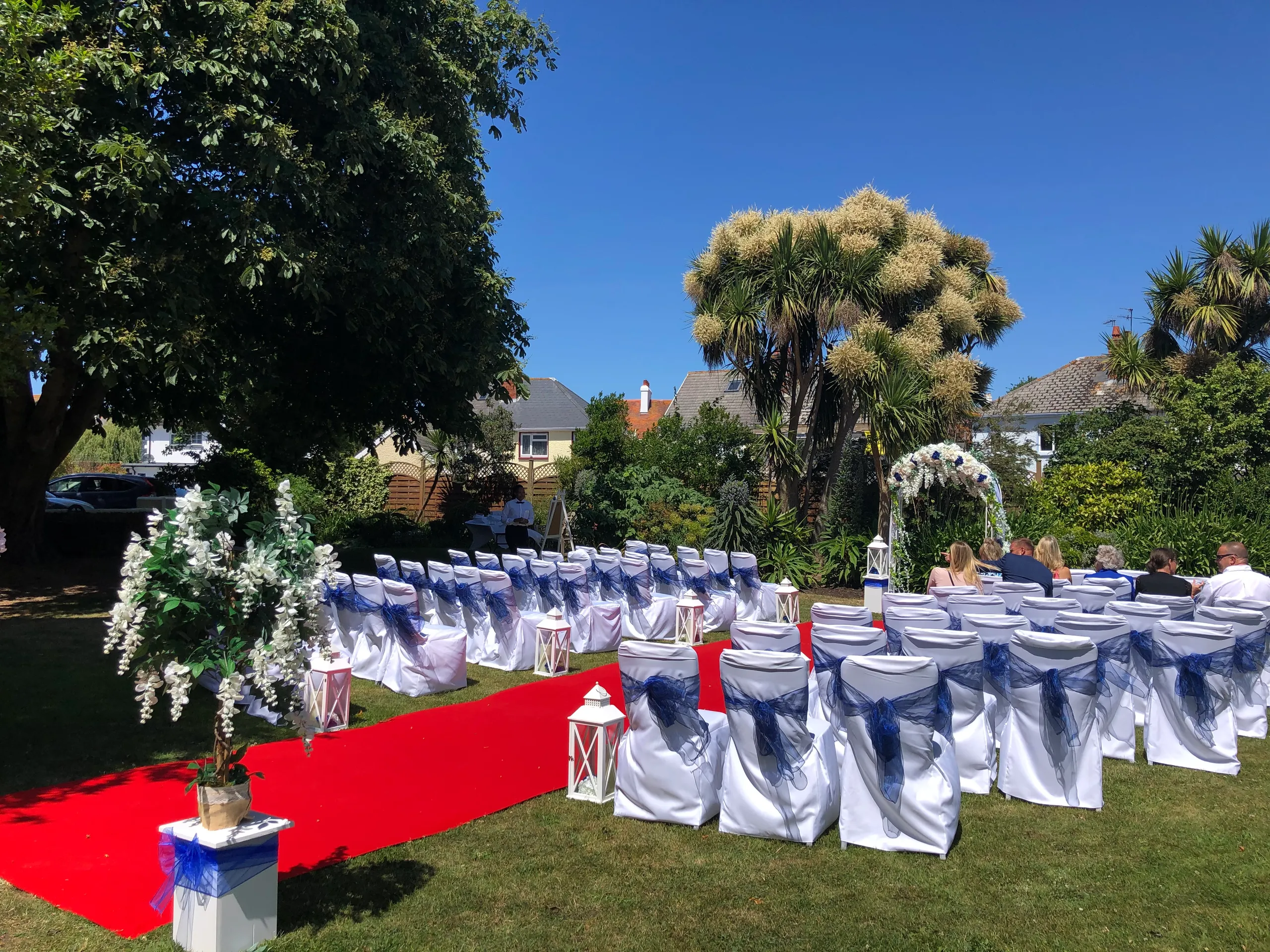 Outdoor wedding ceremony layout in a garden with a red carpet aisle, white chairs tied with blue sashes, floral decorations and a ceremony arch.