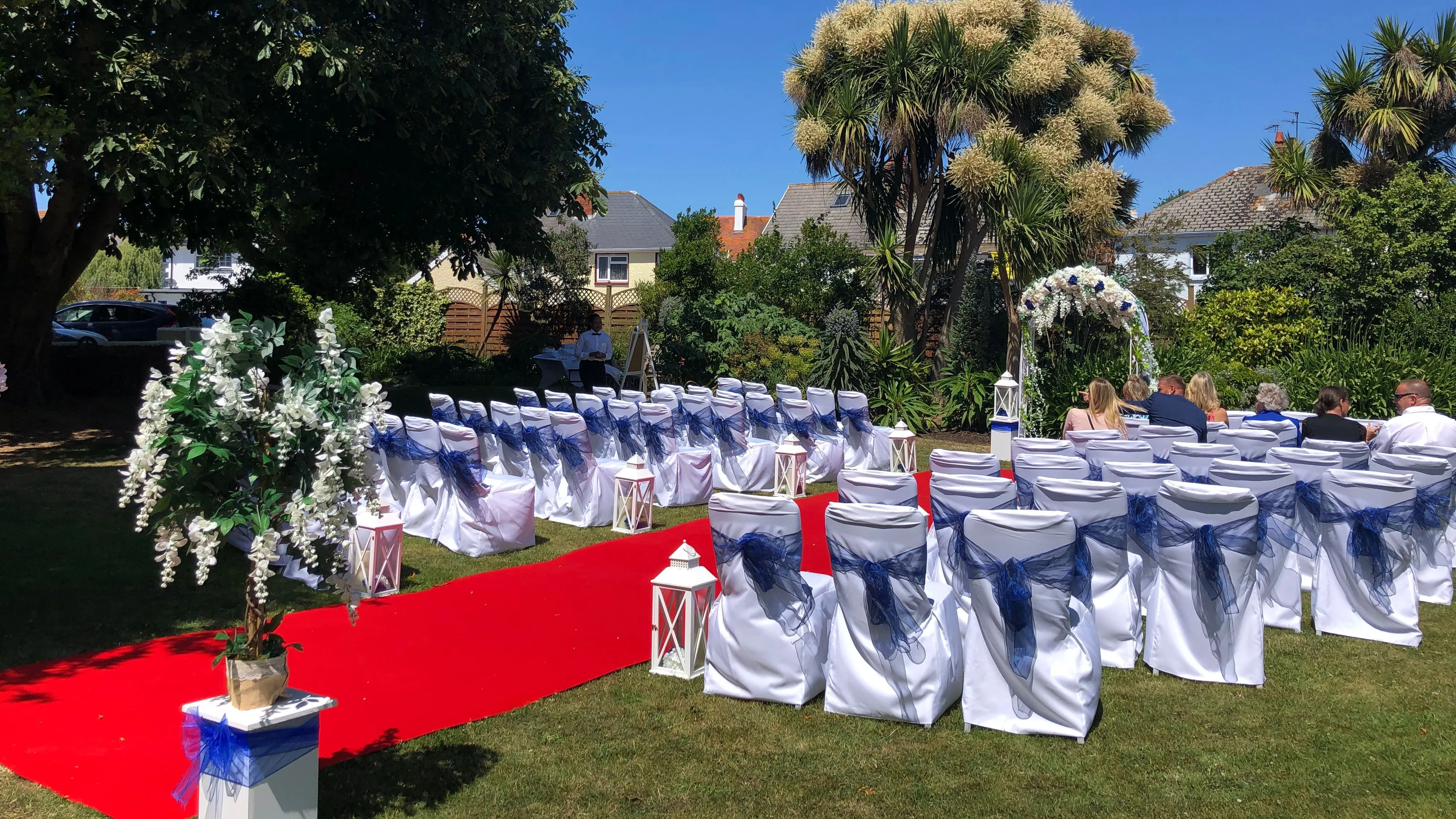 Outdoor wedding ceremony layout in a garden with a red carpet aisle, white chairs tied with blue sashes, floral decorations and a ceremony arch.
