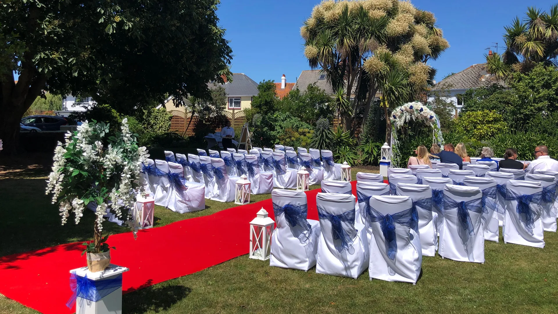 Outdoor wedding ceremony layout in a garden with a red carpet aisle, white chairs tied with blue sashes, floral decorations and a ceremony arch.