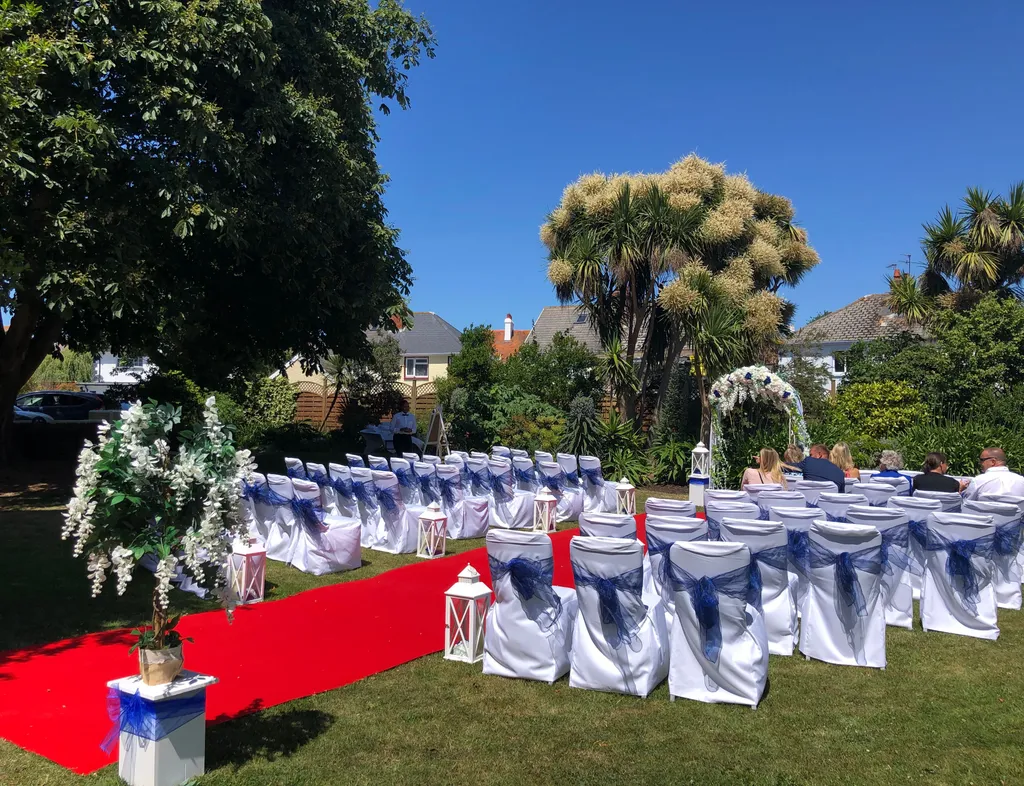 Outdoor wedding ceremony layout in a garden with a red carpet aisle, white chairs tied with blue sashes, floral decorations and a ceremony arch.