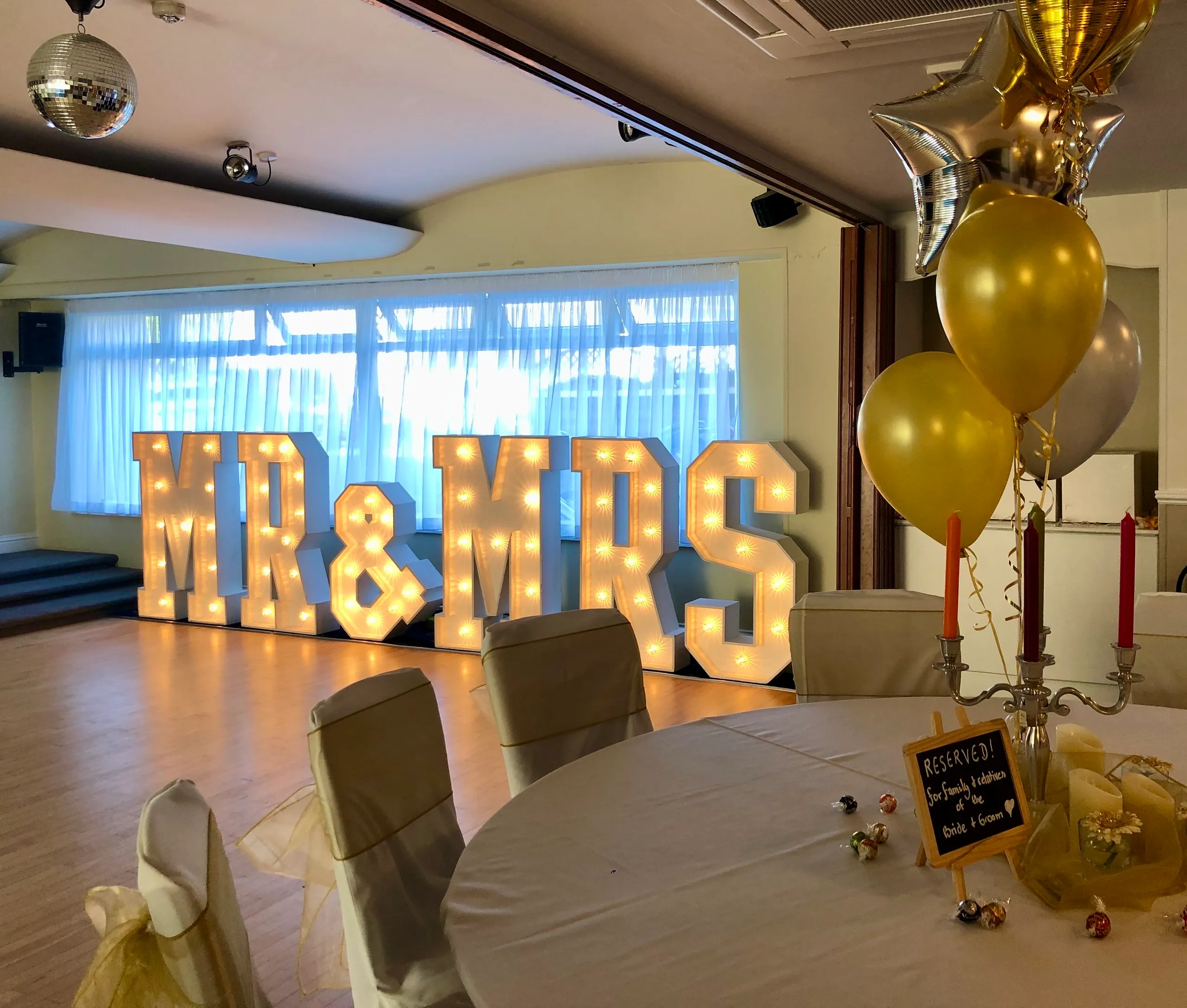 Illuminated Mr & Mrs wedding letters in a decorated reception venue with balloons and table settings