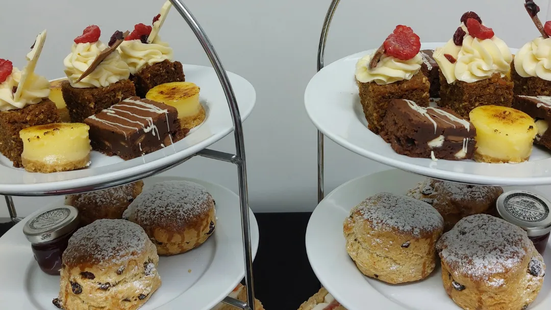 Two-tiered afternoon tea stands with assorted cakes, scones with jam, and finger sandwiches on white plates.