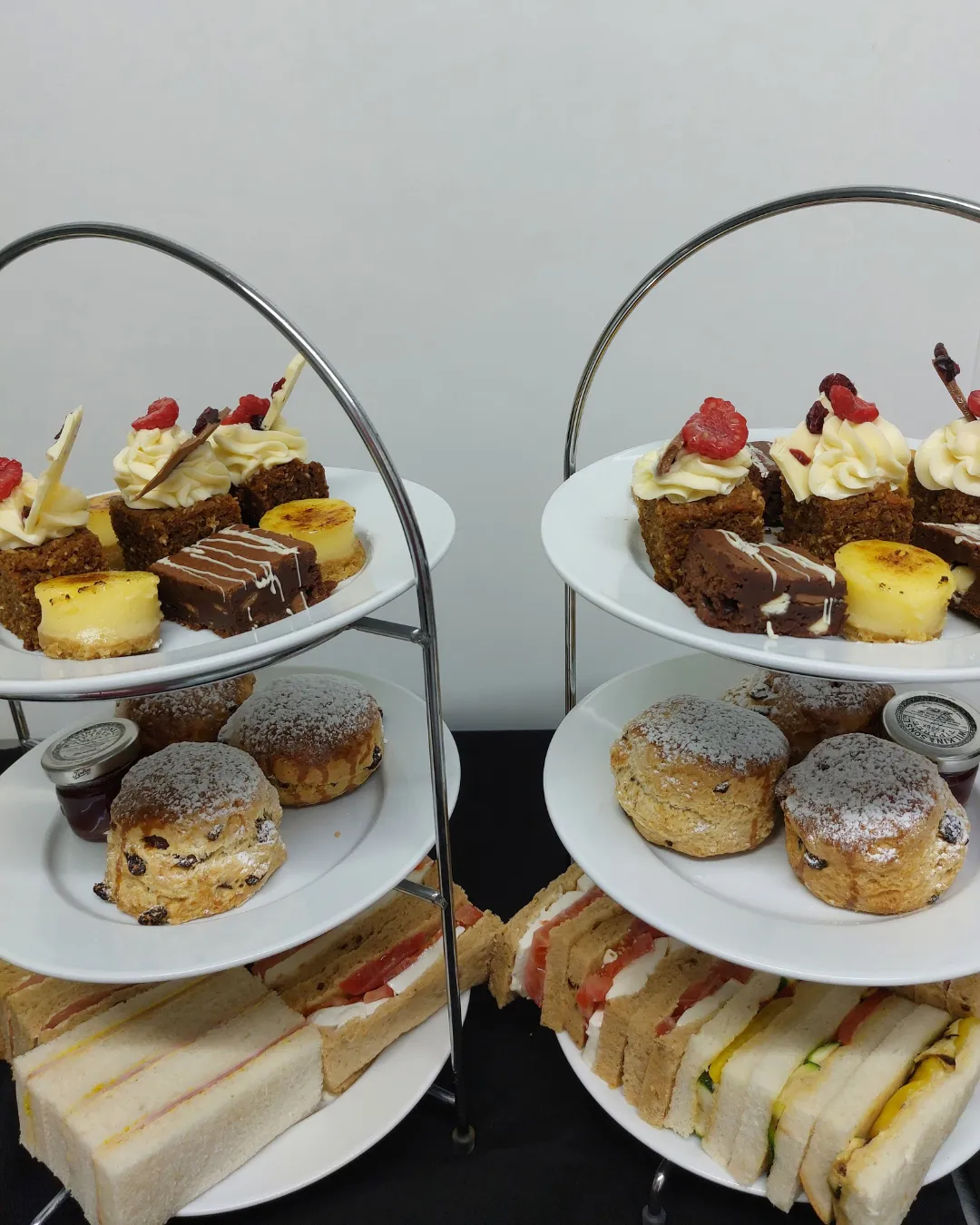 Two-tiered afternoon tea stands with assorted cakes, scones with jam, and finger sandwiches on white plates.