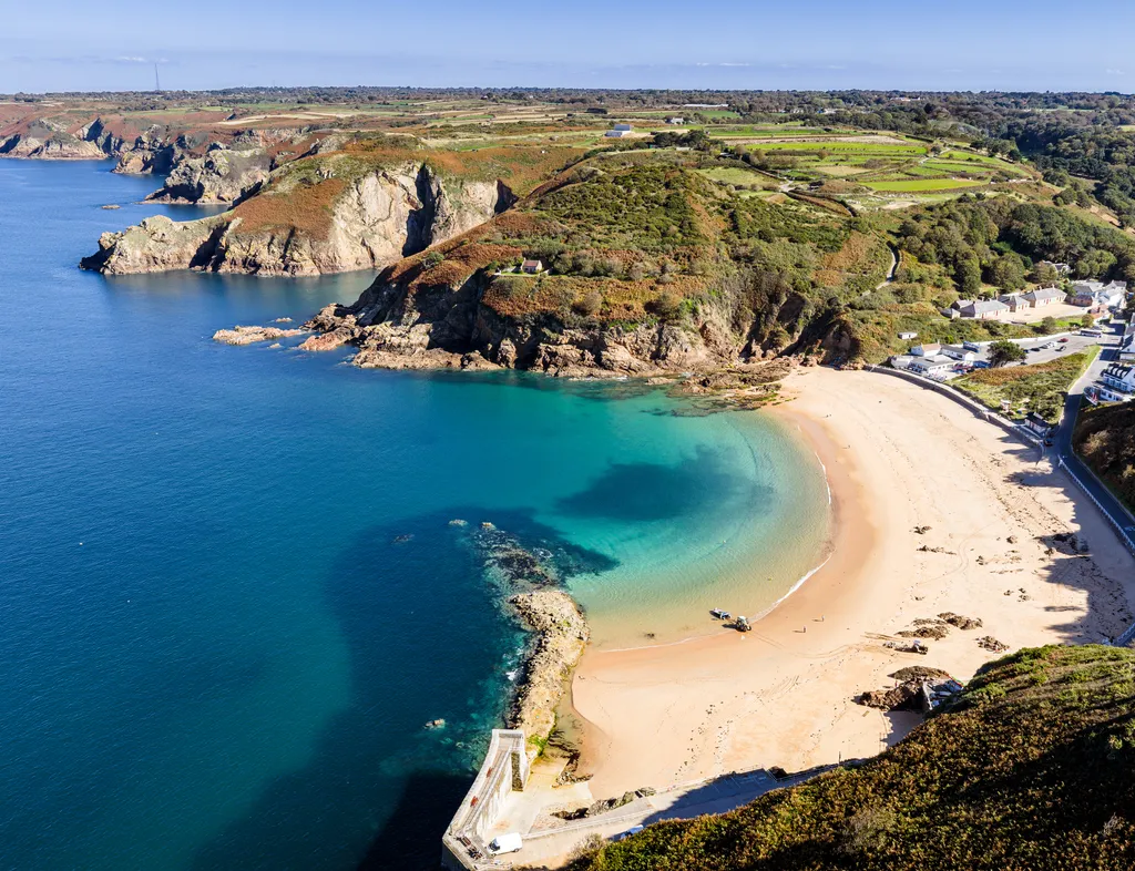 Aerial view of a sandy bay with clear turquoise water, rugged cliffs and a small coastal village.