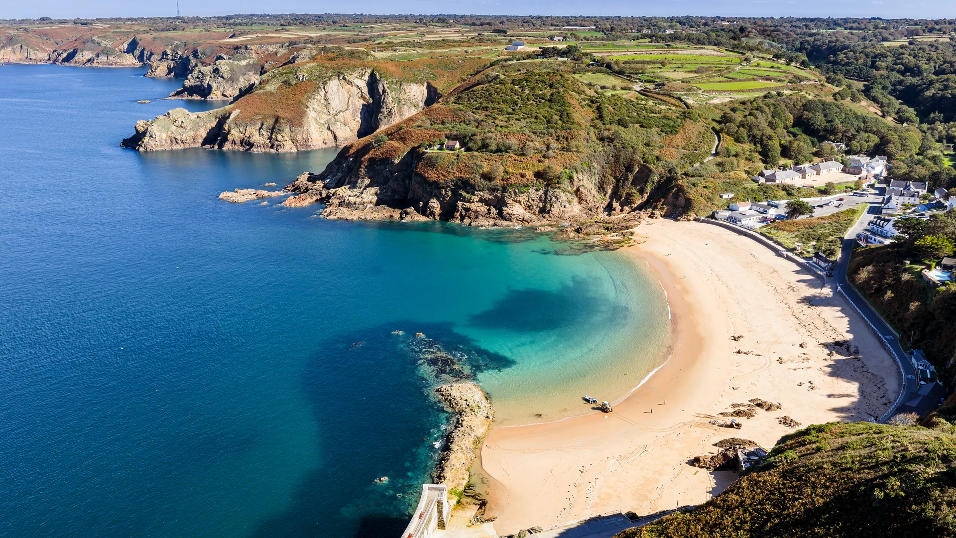 Aerial view of a sandy bay with clear turquoise water, rugged cliffs and a small coastal village.