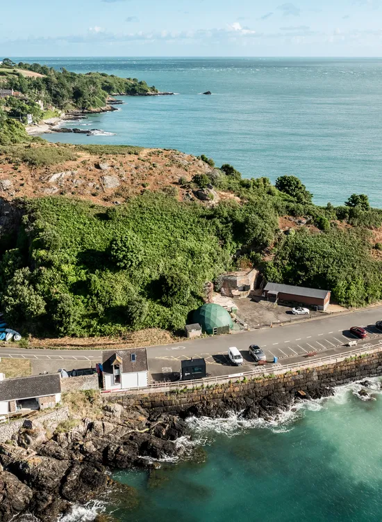 Aerial view of a rocky coastline with turquoise sea, a winding coastal road, and a tree-covered headland.