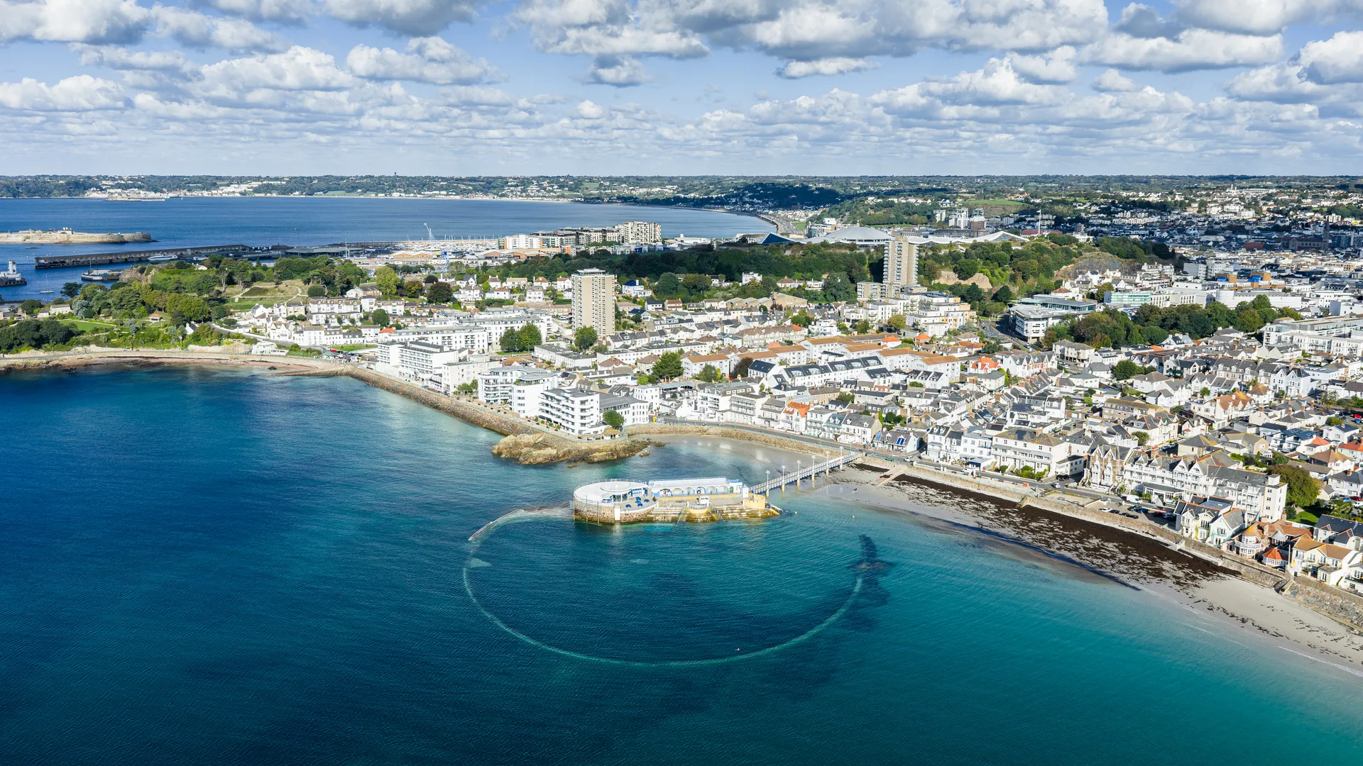 Aerial view of a coastal town with a seaside lido, sandy beach and turquoise bay under a sky filled with scattered clouds.
