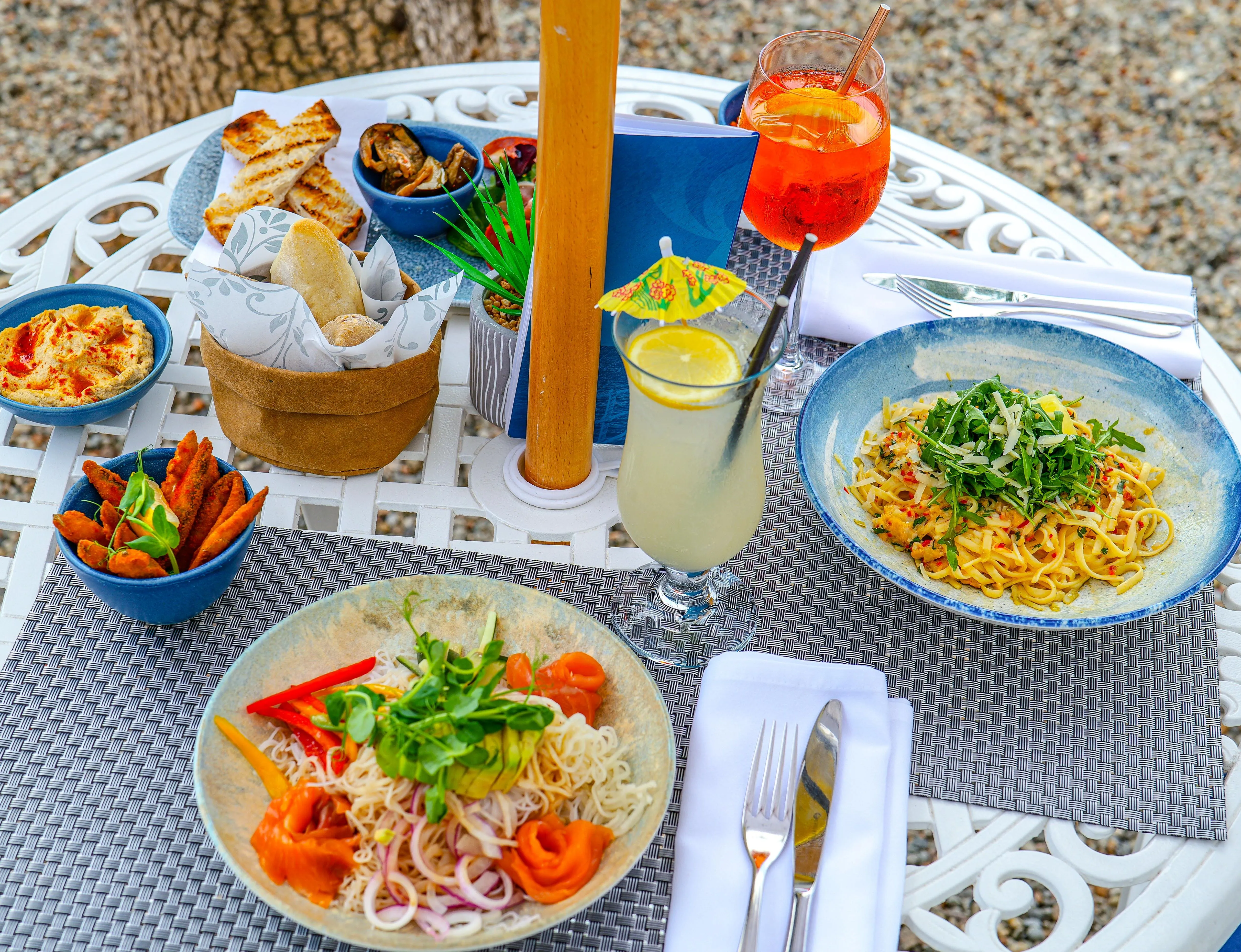 Outdoor terrace dining table with pasta, smoked salmon noodle salad, bread, dips, sweet potato wedges and cocktails.