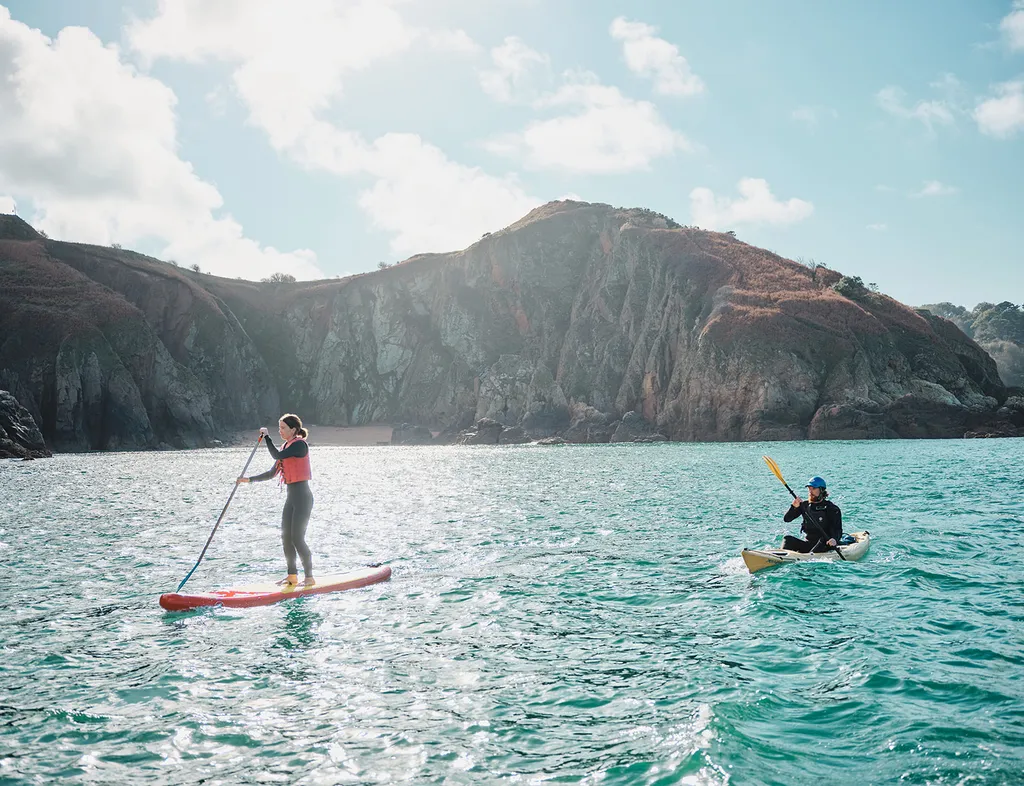 Woman paddleboarding and man kayaking on turquoise sea near rocky cliffs
