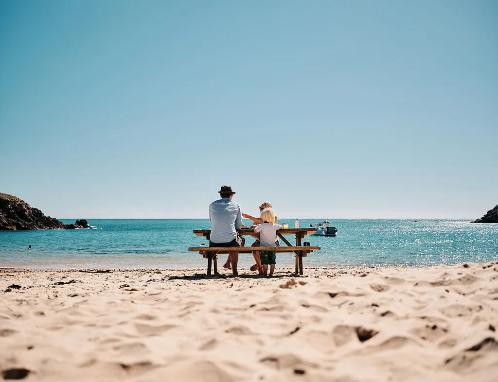 Family sitting at a picnic bench on a sandy beach beside a calm blue sea