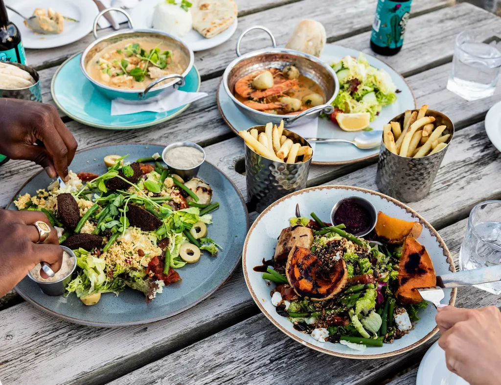 Outdoor dining table with shared plates of salads, seafood curry, chips and flatbread, with people eating together.