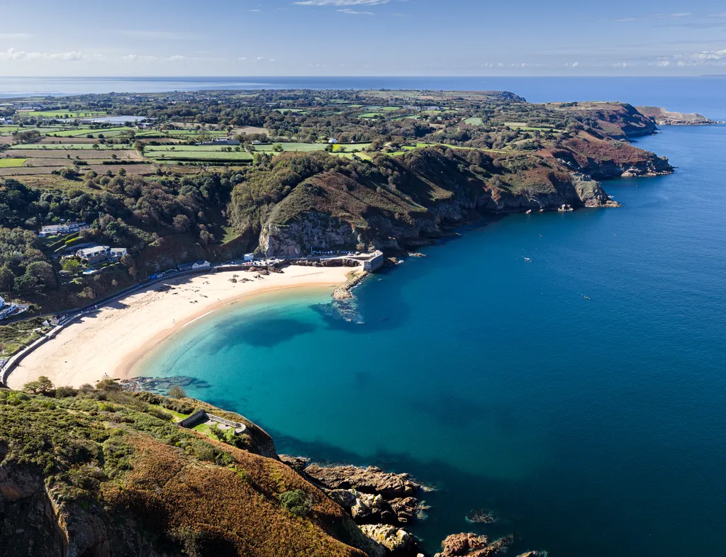 Aerial view of a sweeping sandy bay with turquoise water, rugged cliffs and patchwork countryside beyond.