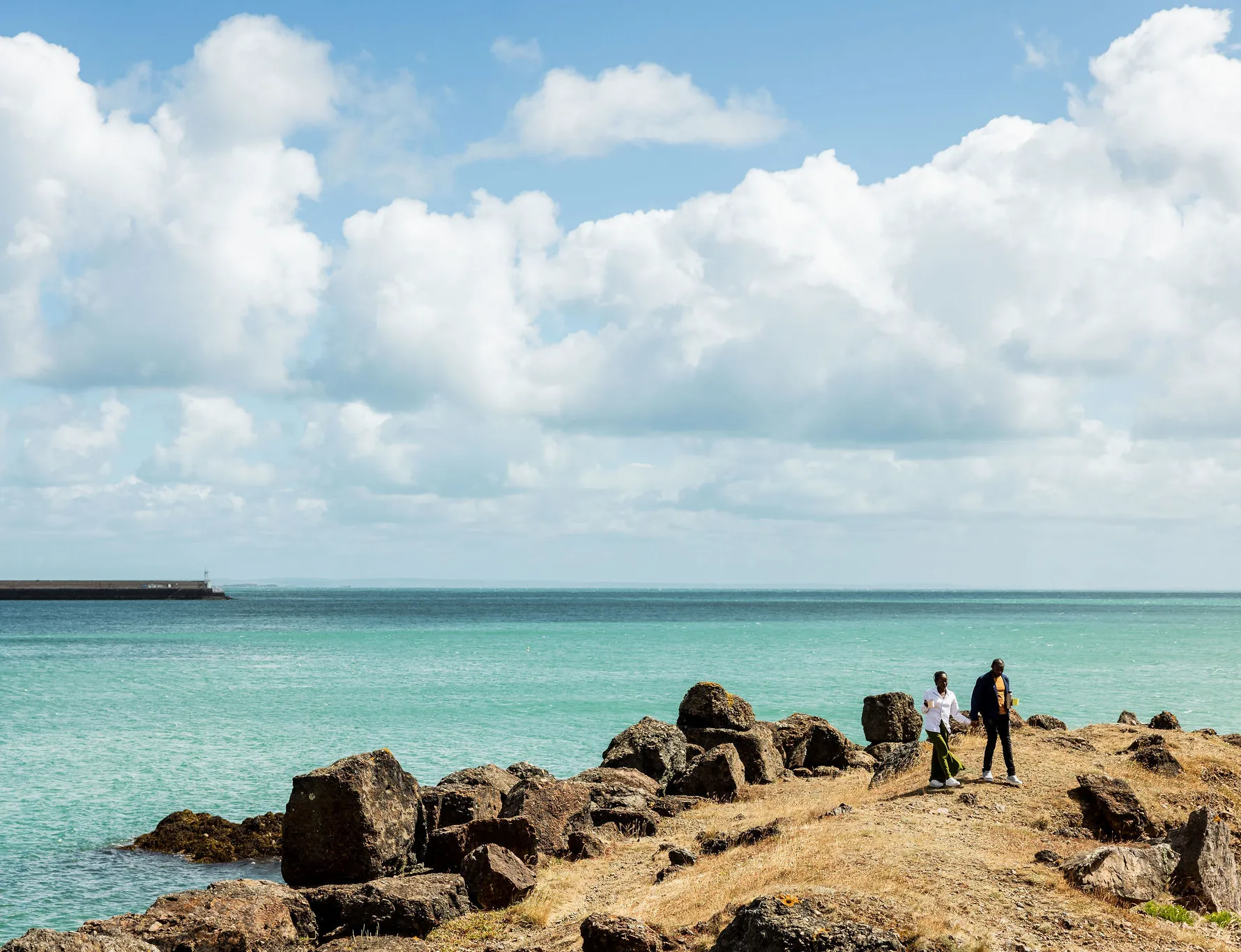 Two people walking along a rocky coastline beside turquoise sea under large white clouds.